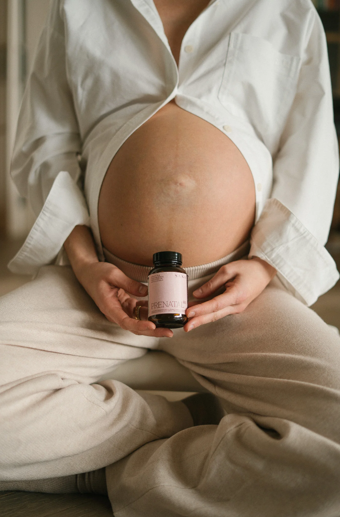 Mujer embarazada sentada sosteniendo un suplemento prenatal SANA Nutrients en las manos, mostrando el abdomen durante el embarazo
