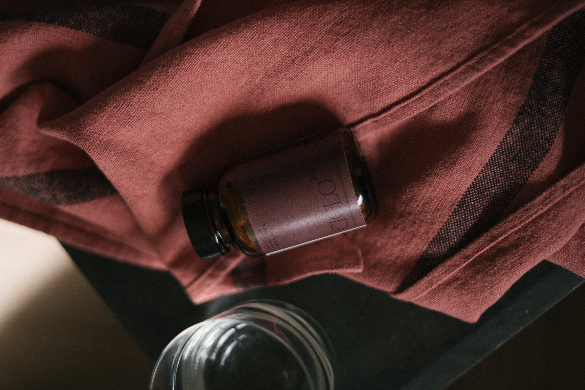 Frasco de Ciclo 360 de SANA sobre tela burdeos, suplemento natural para el ciclo menstrual y bienestar femenino