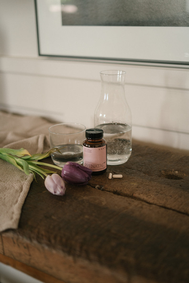 Suplemento prenatal Prenatal 360 de SANA Nutrients con cápsulas junto a vaso de agua en entorno natural para embarazo y fertilidad
